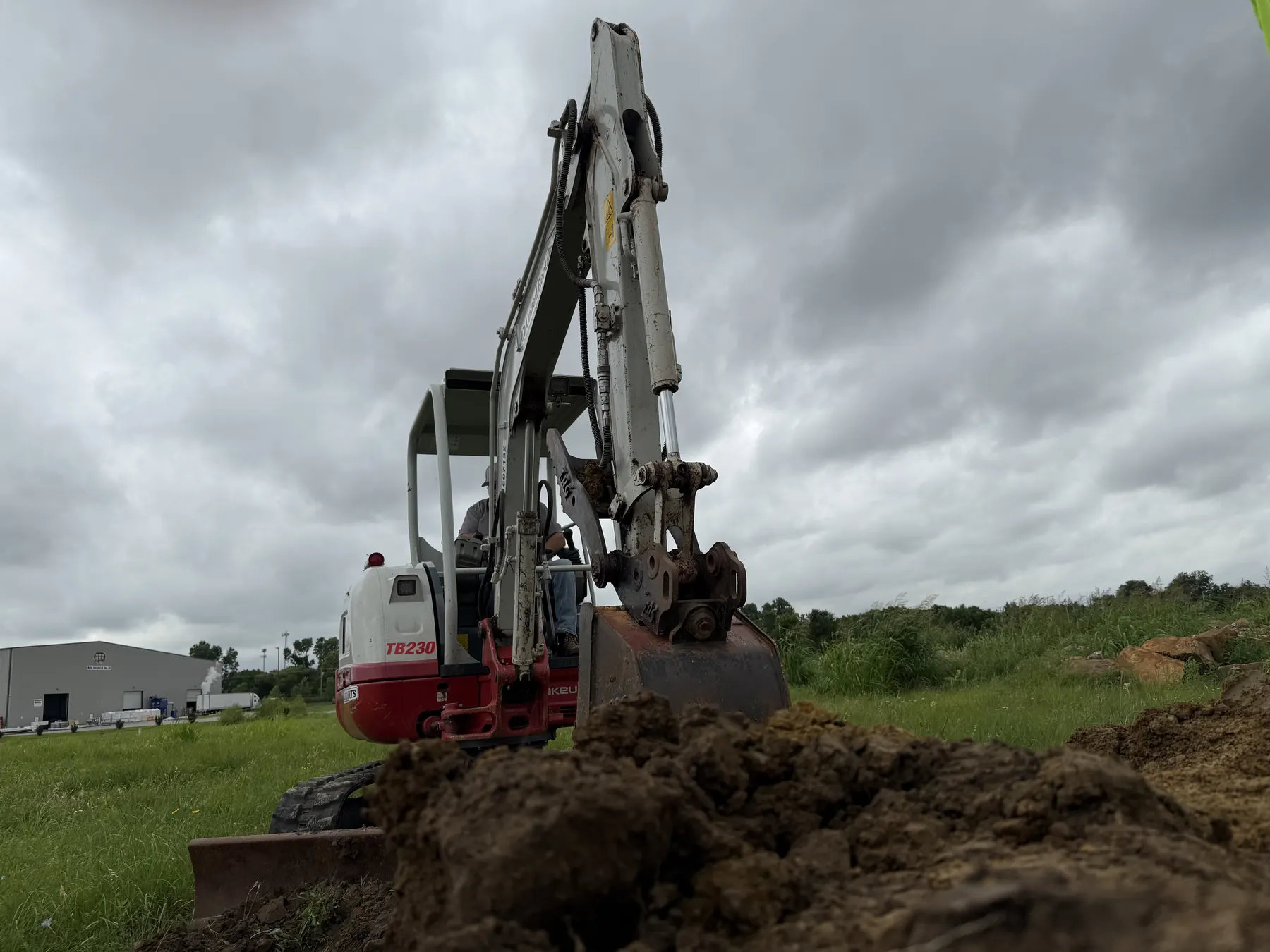 A compact excavator works beside a grassy field with dark soil in the foreground